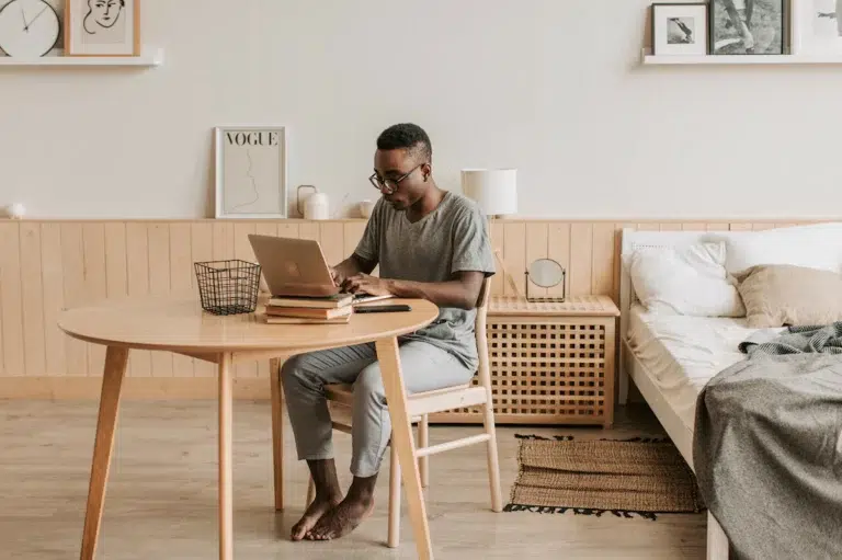 Jeune homme assis à sa table de salon près de son lit travaillant sur son ordinateur portable
