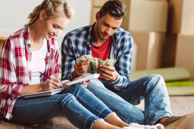 Couple assis au sol au milieu de cartons de déménagement. La femme souriante écrit sur un carnet d'une main et tient une calculatrice de l'autre tandis qu'un homme compte des billets