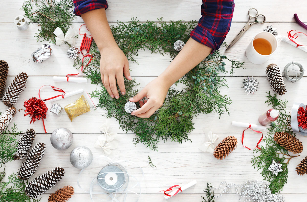 Une personne composant une couronne de Noël avec des boules de Noël, du fil, des pommes de pin et des ciseaux