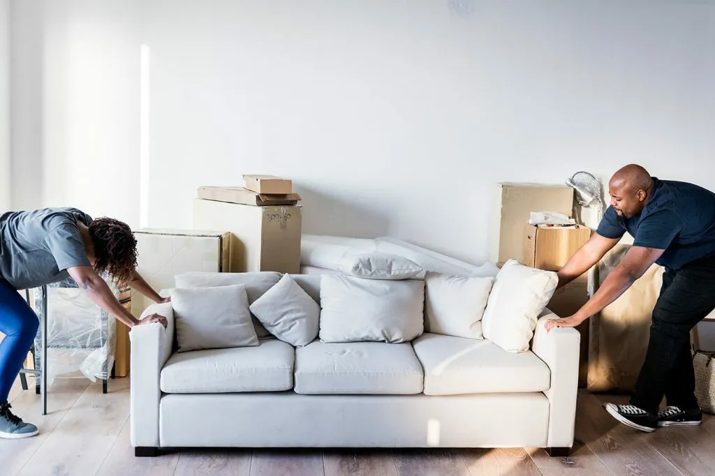 Un couple en plein déménagement essaie de déplacer un canapé beige. La femme le pousse à une extrémité tandis que l'homme le tire à l'autre