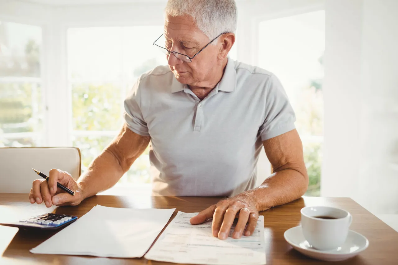 Homme âgé assis à une table, tapant sur une calculatrice devant plusieurs documents éparpillés et une tasse à café