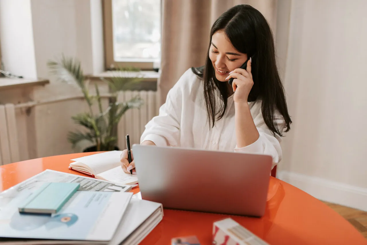 Jeune femme au téléphone prenant des notes sur un carnet devant son ordinateur portable