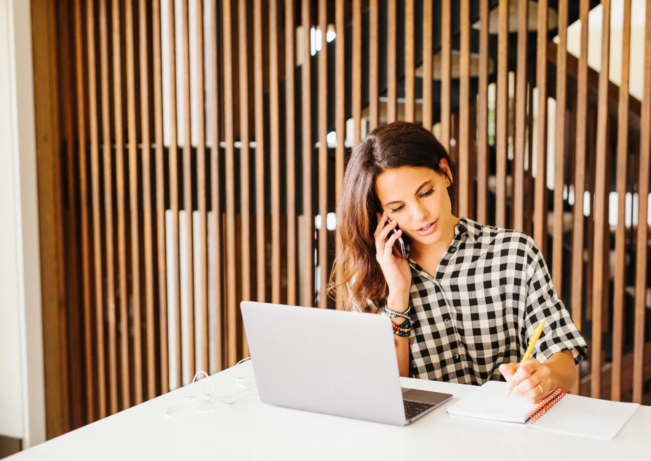 Femme au téléphone devant son ordinateur portable notant sur son petit carnet