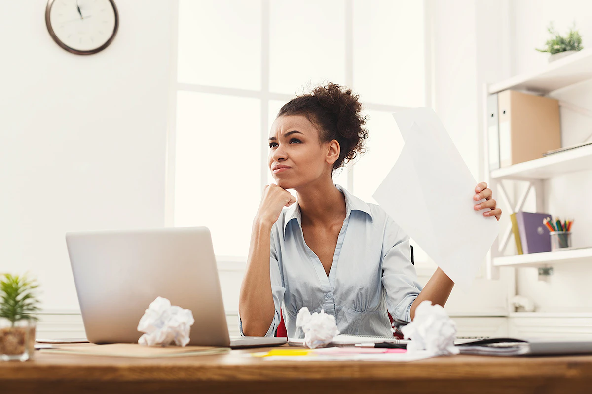 Femme perplexe assise à son bureau plein de papiers chiffonnés, un document à la main