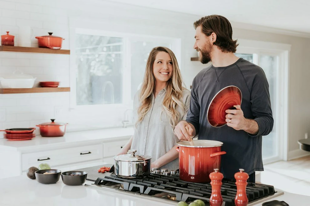 Un couple cuisine un repas dans une casserole rouge au milieu d'une cuisine spacieuse blanche