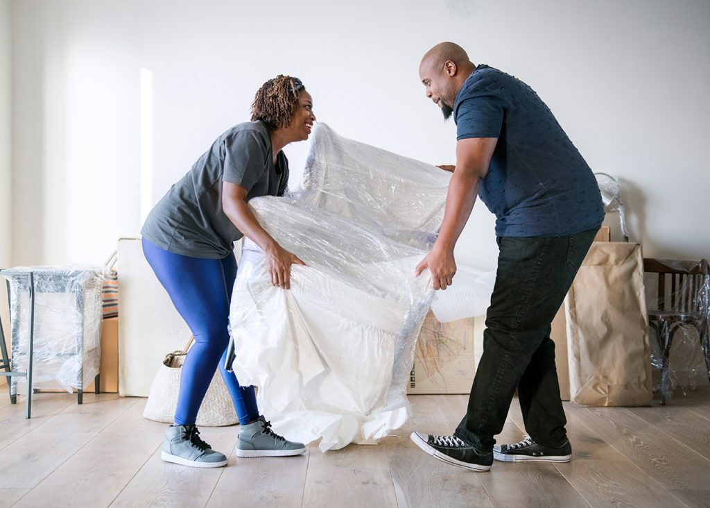 Un homme et une femme qui soulève un fauteuil emballé au milieu d'une pièce remplie de carton de déménagement et de mobilier emballé