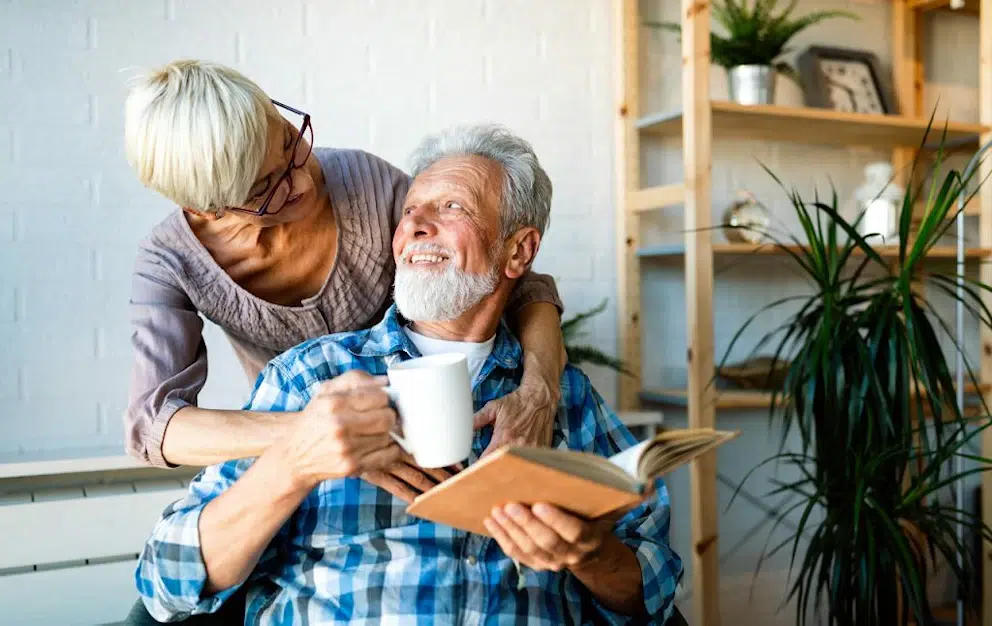 Une femme âgée enlace son mari qui lit un livre et tient une tasse à café