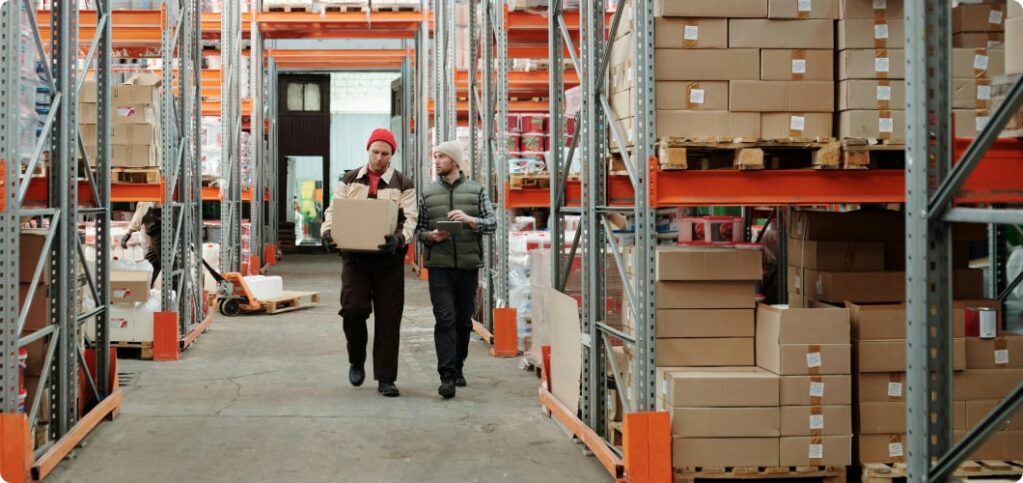 2 hommes marchent dans l'allée principale d'un entrepôt de stockage. L'un d'eux porte un carton.