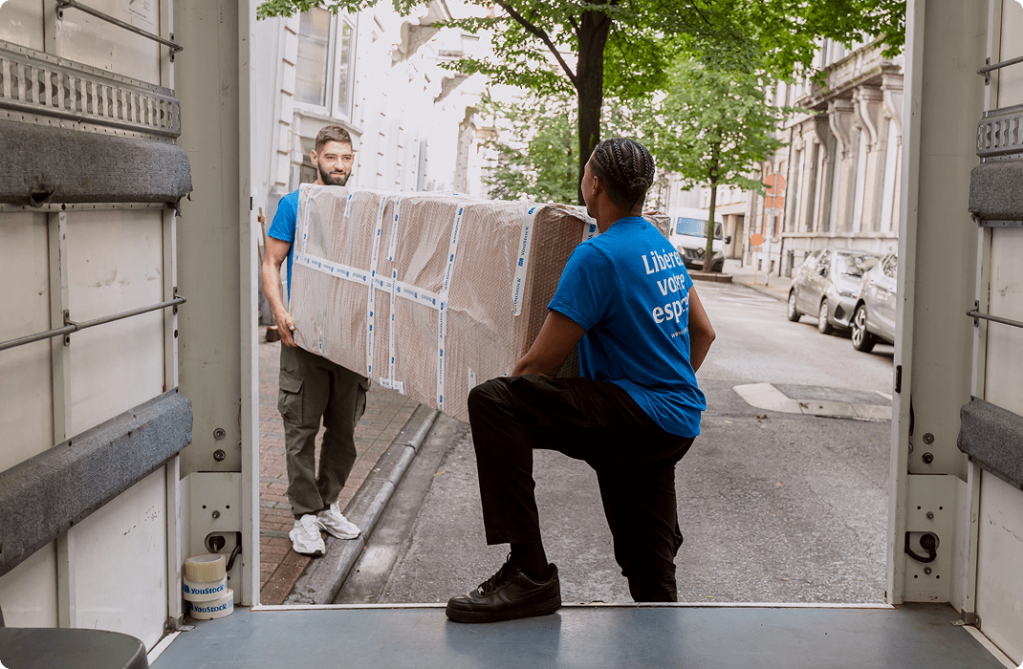 Two removal men carry a wrapped and protected piece of furniture onto a removal truck.