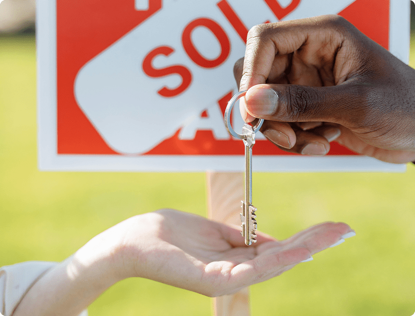 One hand holding a key to another in front of a house for sale sign crossed out with the message ‘sold’.