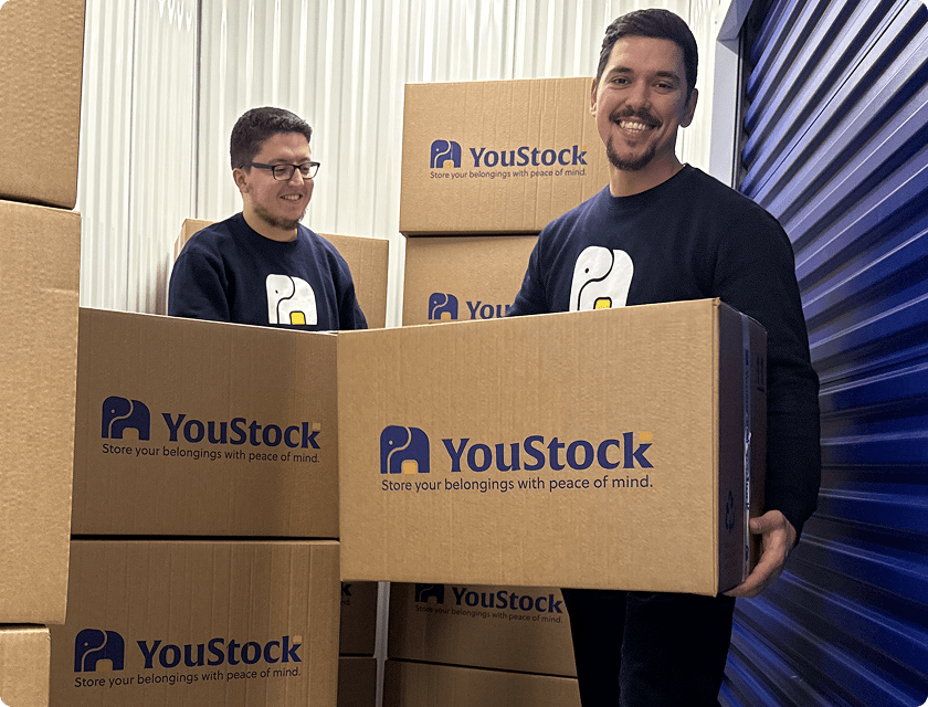 A YouStock mover holds a moving box in the middle of a storage centre aisle filled with YouStock branded boxes.