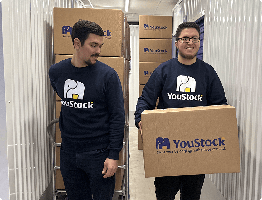 Two YouStock professionals carry cardboard boxes down the aisle of a self-storage centre.