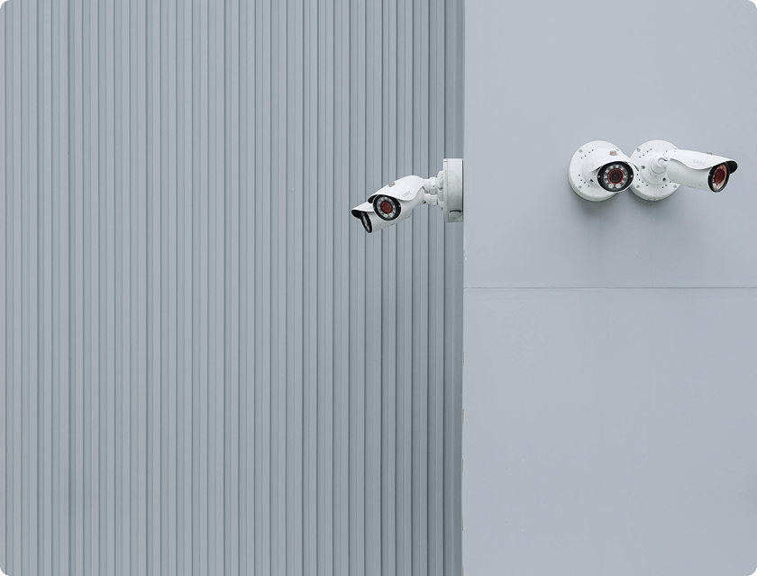 Zoom in on 4 video surveillance cameras in the corner of a grey warehouse.