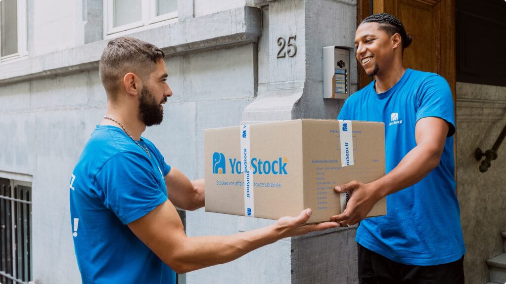 Two YouStock movers pass around a storage and removal box at the bottom of a building.