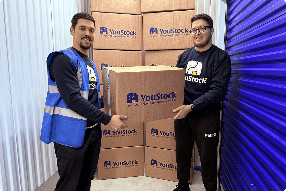 Two storage professionals carry a box in front of a stack of YouStock branded moving boxes.