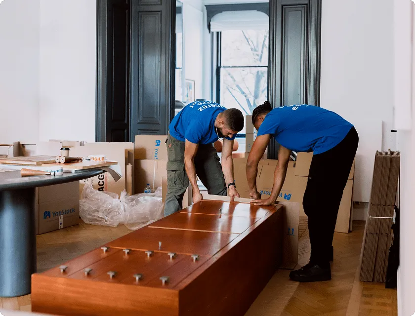 Two YouStock removal men pass a moving box between them.