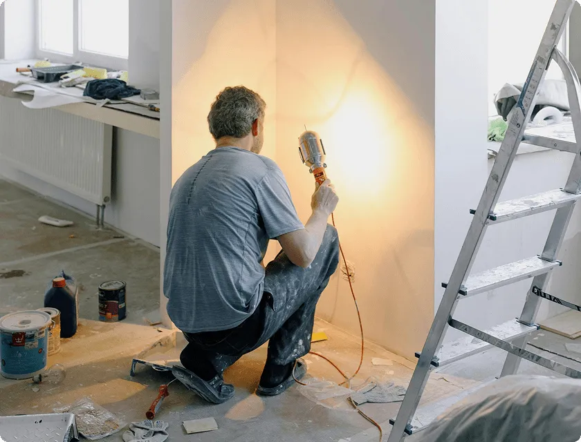 A man crouching on a construction site uses a light to inspect a repainted wall.