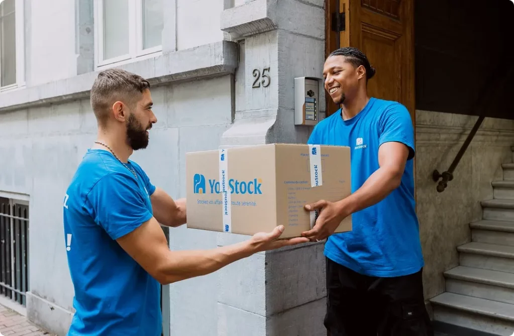 Deux déménageurs YouStock se transmettent un carton de stockage / déménagement devant l'entrée d'un immeuble