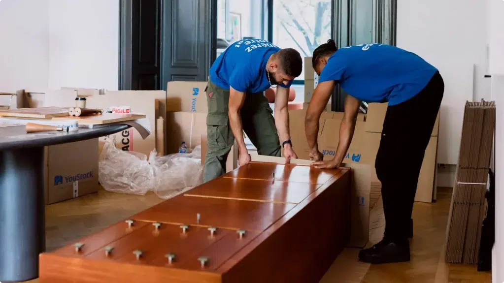 Two YouStock removal men pack and protect a wooden piece of furniture for moving.