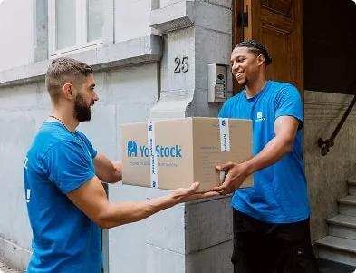 Deux déménageurs YouStock se transmettent un carton de stockage / déménagement devant l'entrée d'un immeuble