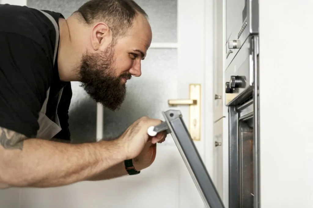 Homme barbu portant un tablier ouvre la porte du four et regarde à l'intérieur