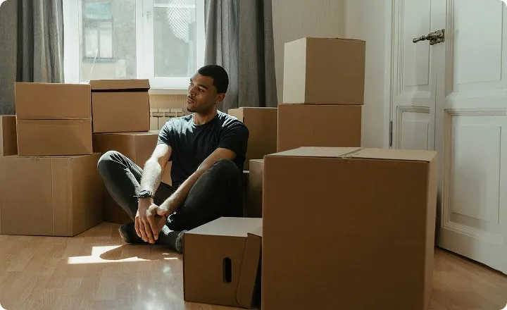 Un jeune homme est assis par terre dans une pièce remplie de cartons de déménagement