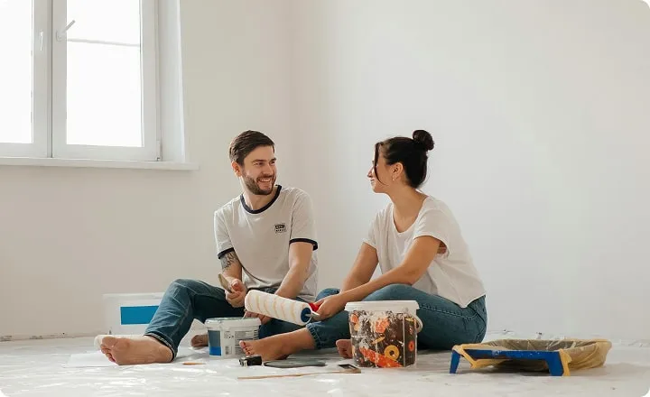 Un couple est assis par terre au milieu d'une pièce vide et de pots de peinture