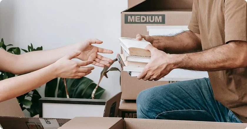 A man hands a stack of books to a woman who holds out her hands. They are in a room full of moving boxes.