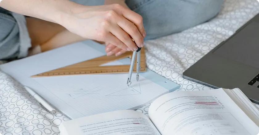 A young student surrounded by a maths book and a laptop draws a geometric figure in a notebook.