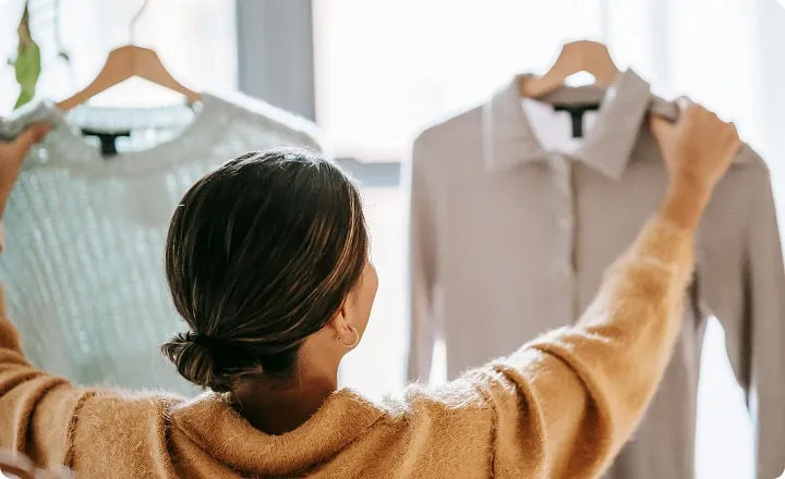 Une jeune femme soulève un pull et une chemise sur cintre devant elle