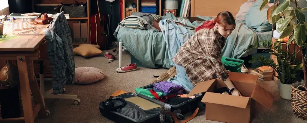 A student is packing a box and her suitcase on the floor of her university room.