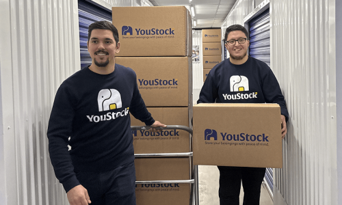 alternative-stackt-storage-service Two YouStock professionals carry cardboard boxes down the aisle of a self-storage centre.