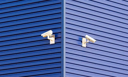 Zoom in on 2 video surveillance cameras in the corner of a blue warehouse.