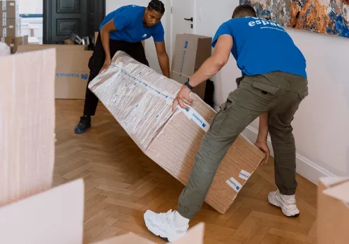 Two YouStock movers return a piece of furniture, packed and protected, in a room filled with storage boxes.